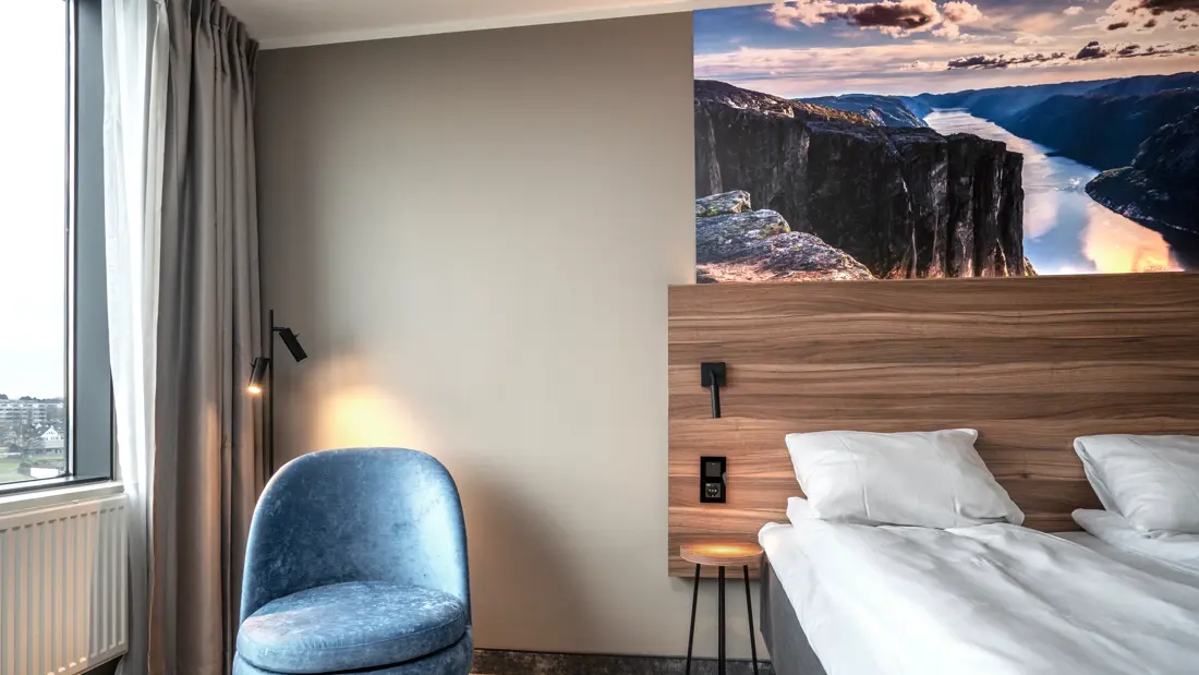 Twinrom med blå hvilestol, gulvlampe, nattbord og to enkeltsenger satt inntil hverandre. Teppebelagt gulv, vindu og panelovn.