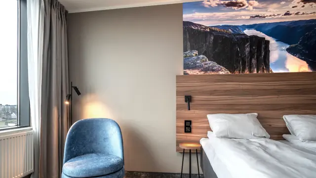 Twinrom med blå hvilestol, gulvlampe, nattbord og to enkeltsenger satt inntil hverandre. Teppebelagt gulv, vindu og panelovn.