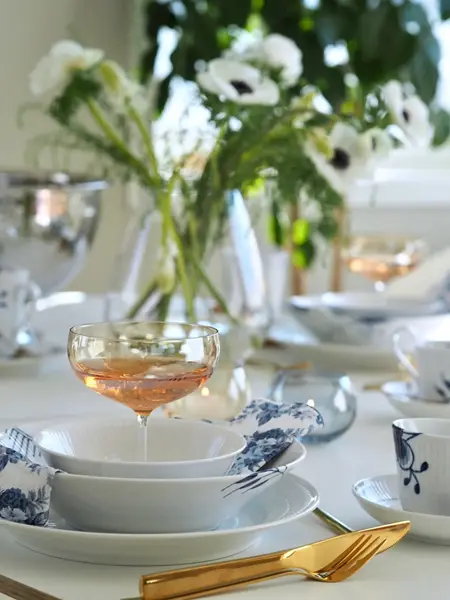 Nærbilde av kuvert med gullbestikk, blått og hvitt service og et coctailglass. Vase med hvite blomster i bakgrunnen.