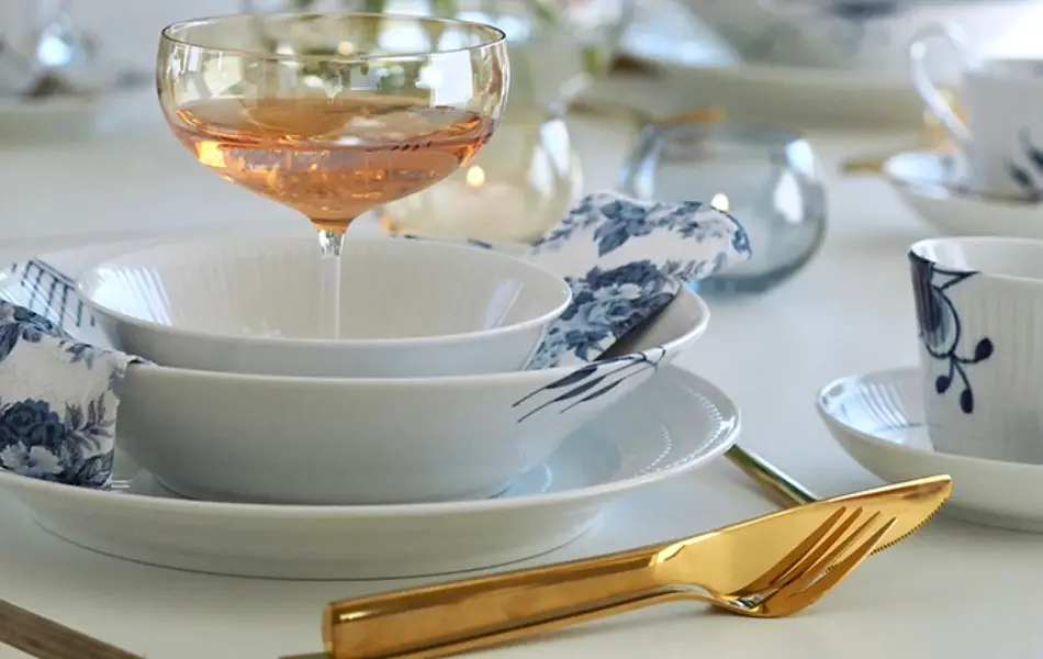 Nærbilde av kuvert med gullbestikk, blått og hvitt service og et coctailglass. Vase med hvite blomster i bakgrunnen.