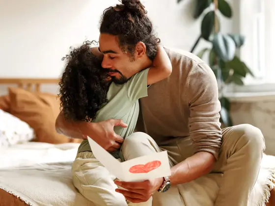 Mann som sitter på seng i et lyst soverom får et kort med hjerte og en klem fra en jente. Foto til artikkel om gaver til farsdag.