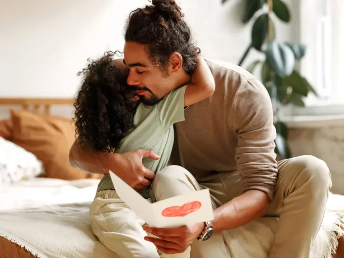 Mann som sitter på seng i et lyst soverom får et kort med hjerte og en klem fra en jente. Foto til artikkel om gaver til farsdag.