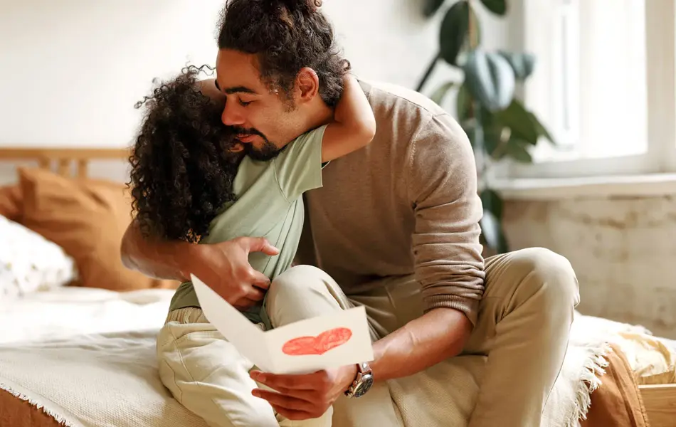 Mann som sitter på seng i et lyst soverom får et kort med hjerte og en klem fra en jente. Foto til artikkel om gaver til farsdag.
