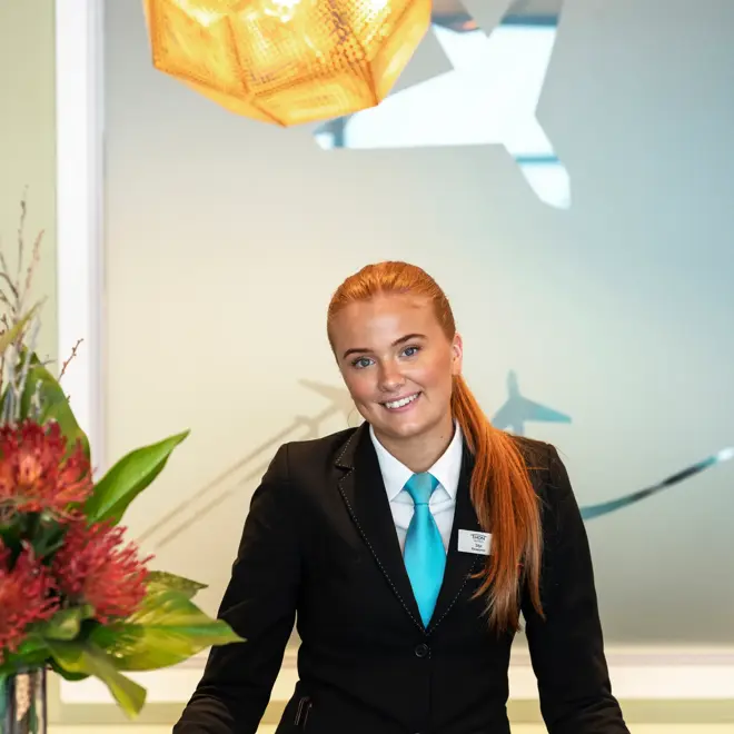 En kvinnelig resepsjonist i uniform stående bak en resepsjonsdisk, blomster i vase. 