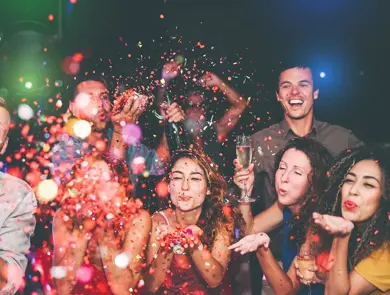 Folkemengde blåser fargerikt konfetti mot kamera. Foto til artikkel om å arrangere nyttårsfest