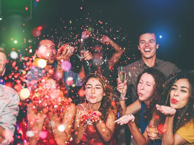 Folkemengde blåser fargerikt konfetti mot kamera. Foto til artikkel om å arrangere nyttårsfest