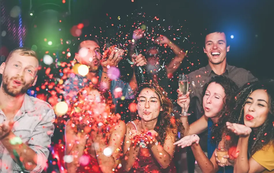 Folkemengde blåser fargerikt konfetti mot kamera. Foto til artikkel om å arrangere nyttårsfest