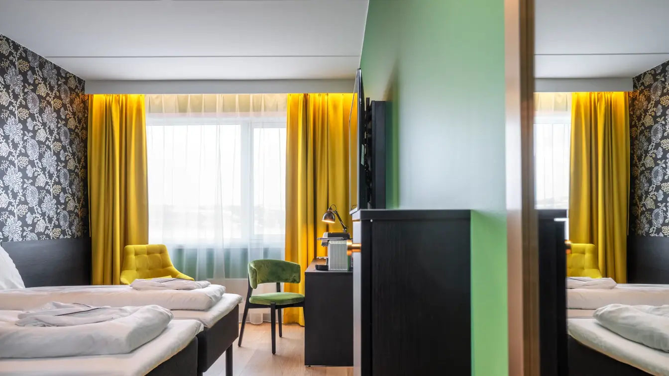 Separate senger, mørk mønstrete tapet med blomster, gule gardiner, gul stol, skrivebord med kaffemaskin og grønn stol i superior twin rom på Thon Hotel Kirkenes