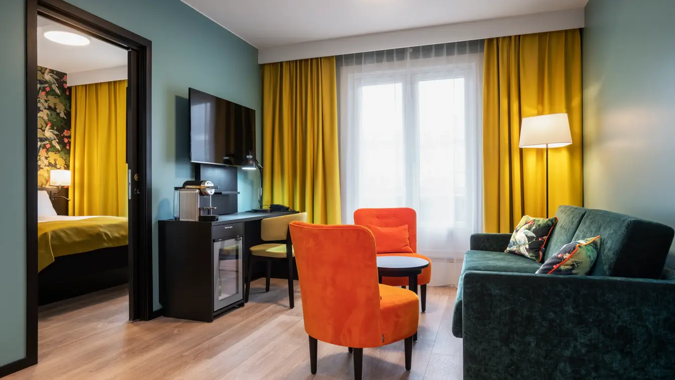 Parkettgulv, vindu fra gulv til tak, gule gardiner, turkise vegger, en grønn sofa, to oransje stoler, flatskjerm hengende på vegg, skrivebord, døråpning med innsyn til et soverom. 