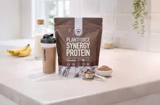 En pose med Synergy Protein fra Plantforce står på en kjøkkenbenk med en shaker med drikke ved siden av.