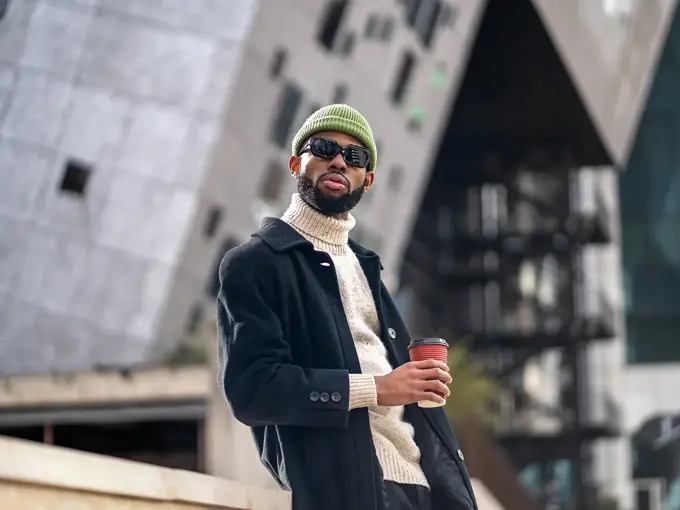 Mann med grønn lue, svart vinterfrakk og solbriller holder kaffekopp 