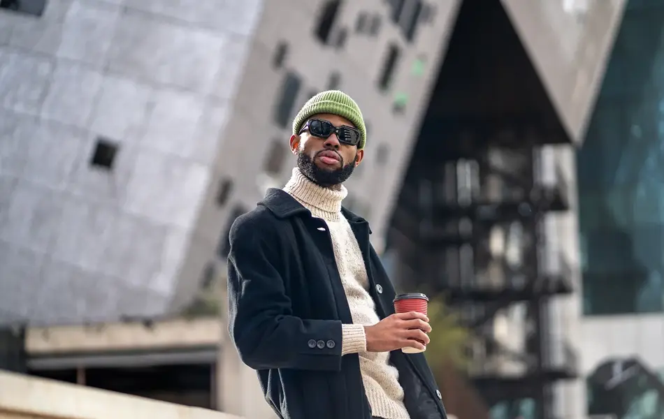 Mann med grønn lue, svart vinterfrakk og solbriller holder kaffekopp 