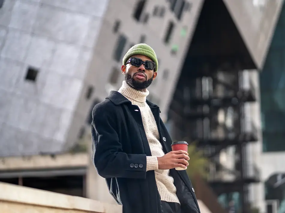 Mann med grønn lue, svart vinterfrakk og solbriller holder kaffekopp 