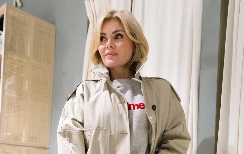 Dame i jeans, collegegenser og beige trenchcoat. Holder en sort veske i hånden