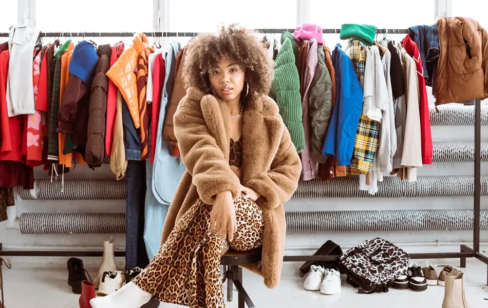 Dame med brun fluffy ullkåpe og leopardmønstret bukse sitter på en stol og ser mot kamera, med klesstativ med fargerike i klær i bakgrunnen.