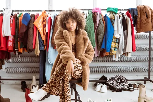 Dame med brun fluffy ullkåpe og leopardmønstret bukse sitter på en stol og ser mot kamera, med klesstativ med fargerike i klær i bakgrunnen.