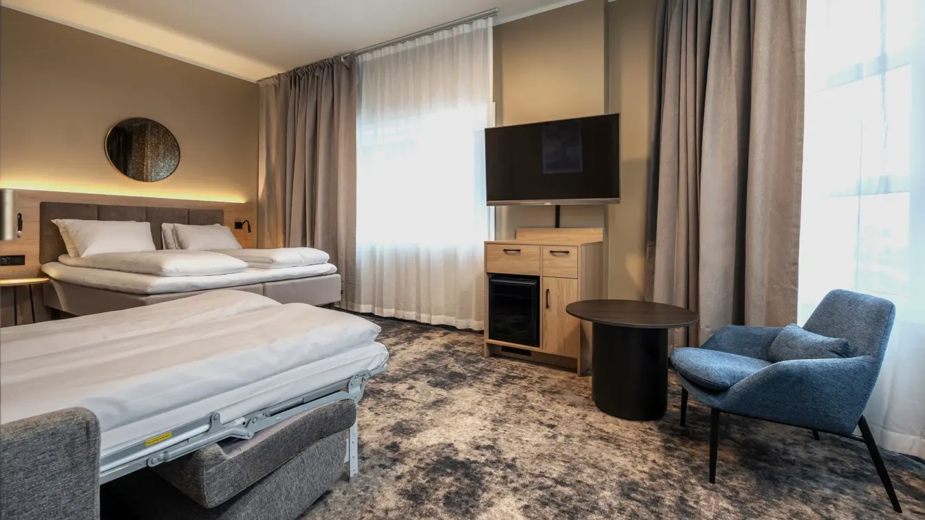 Junior suite med dobbeltseng og oppredd sovesofa, blå lenestol og sort bord, TV og TV benk under med minibar.