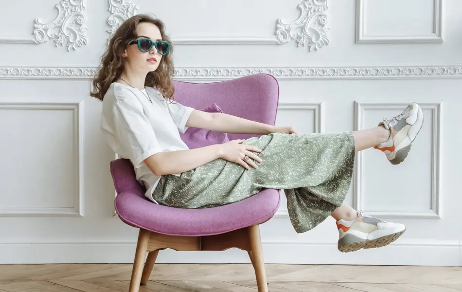 En kvinne halvveis ligger i en lilla stol i et rom med parkett og hvitt panel. Hun har sneakers på føttene, lyse klær og grønne solbriller med hjerter.