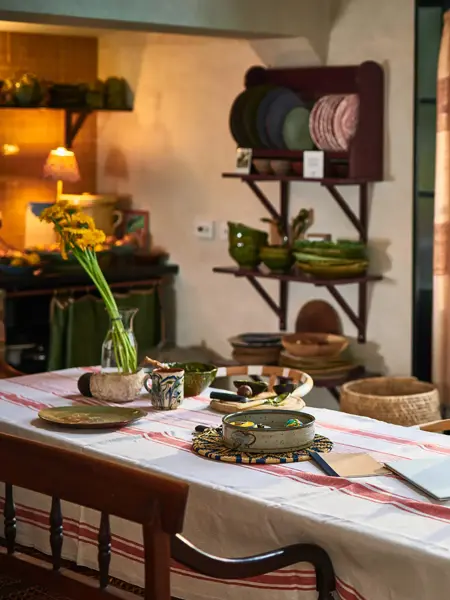 Et spisebord foran en verandadør med hvit og rød duk, dekket med diverse keramikkgjenstander og en vase med blomsterstilker.