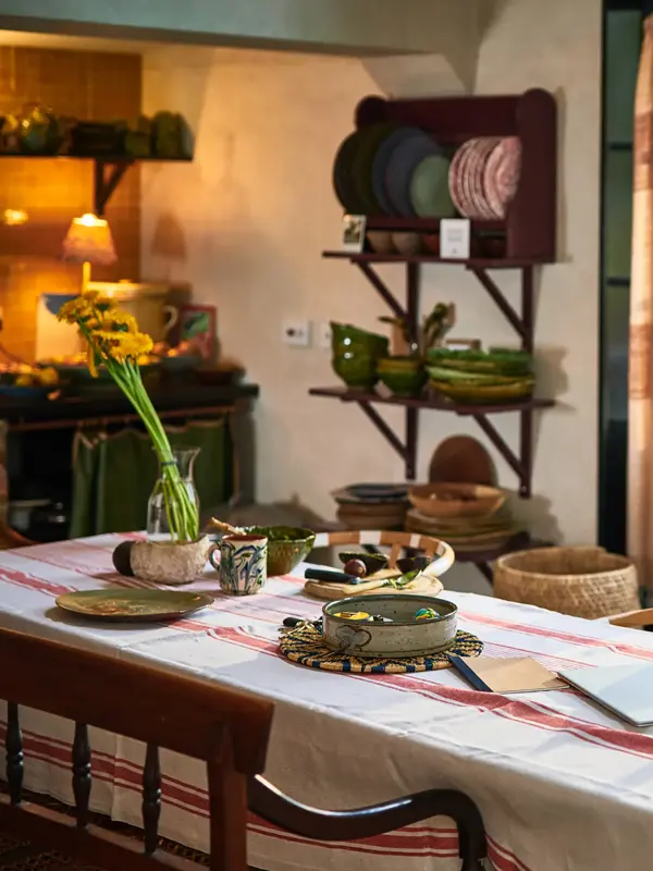 Et spisebord foran en verandadør med hvit og rød duk, dekket med diverse keramikkgjenstander og en vase med blomsterstilker.
