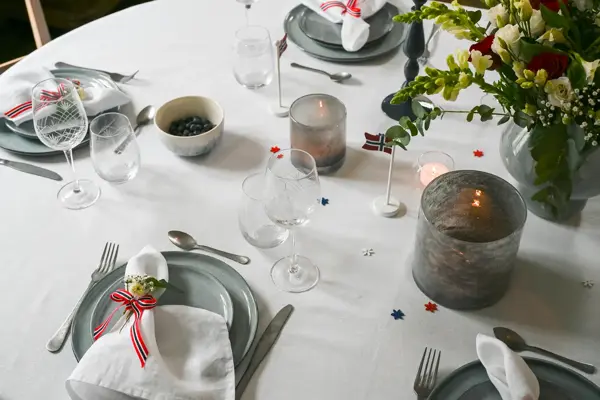 Bord dekket med hvit duk, grå vase, grå telysholder, grått service, hvit linserviett bundet sammen med flaggbånd og blomsterpynt, vinglass og vannglass.