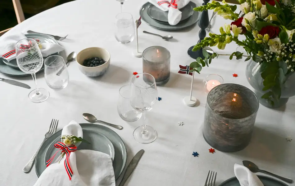 Bord dekket med hvit duk, grå vase, grå telysholder, grått service, hvit linserviett bundet sammen med flaggbånd og blomsterpynt, vinglass og vannglass.