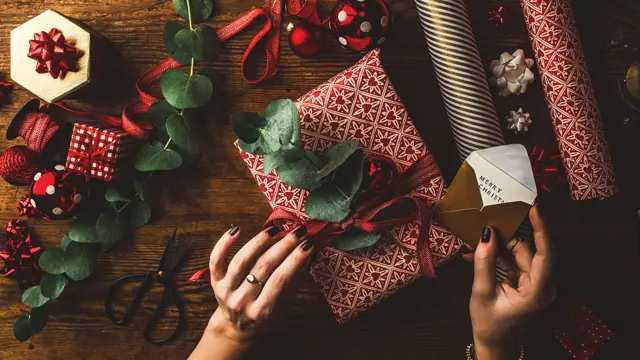 Woman packing contemporary christmas gifts, on table with red rustic paper and green leaves.