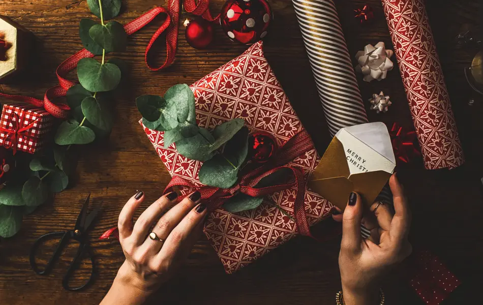 Woman packing contemporary christmas gifts, on table with red rustic paper and green leaves.
