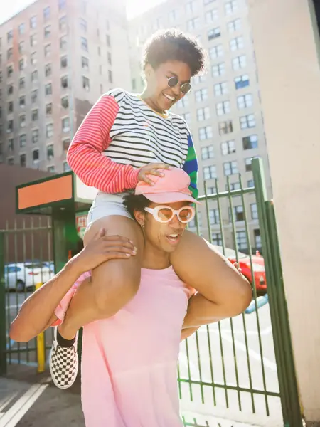 Ung mann iført rosa t-skjorte, solbriller og caps med en ung dame på skuldrene.  