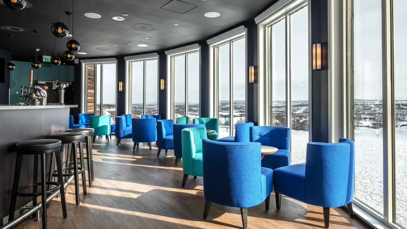 Bar område med mange sitteplasser langs store vinduer med utsikt. Alle lenestolene er blå og turkise og står rundt runde små bord. Bardisk på venstre side. 