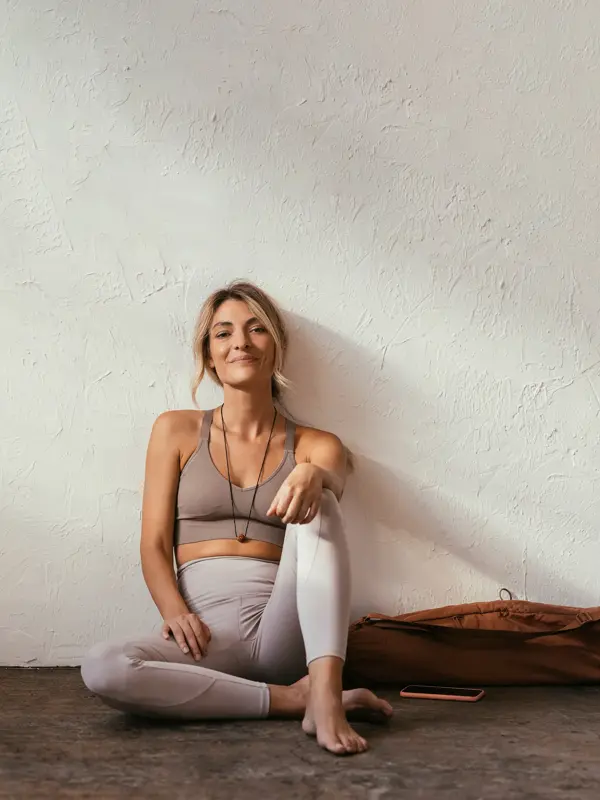 Dame med beige tights og beige sports BH poserer sittende inntil hvit murvegg 