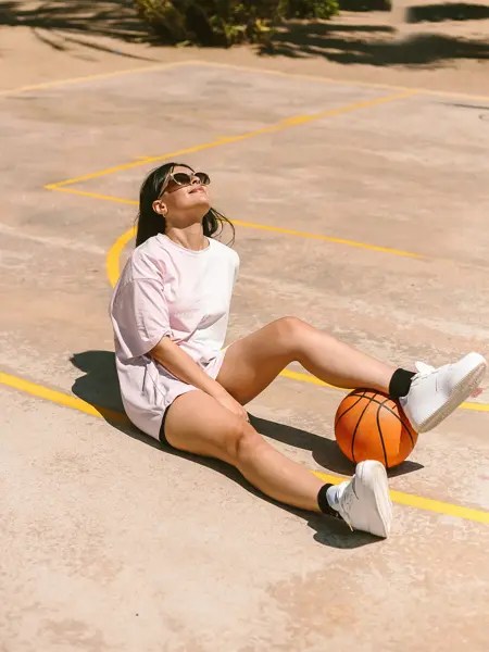 Jente med hvit t-skjorte og sporty shorts sitter på bakken på en basketballbane med benet over en basketball