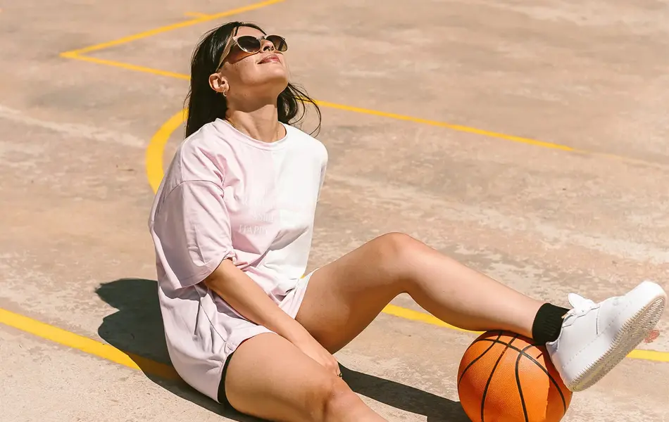 Jente med hvit t-skjorte og sporty shorts sitter på bakken på en basketballbane med benet over en basketball