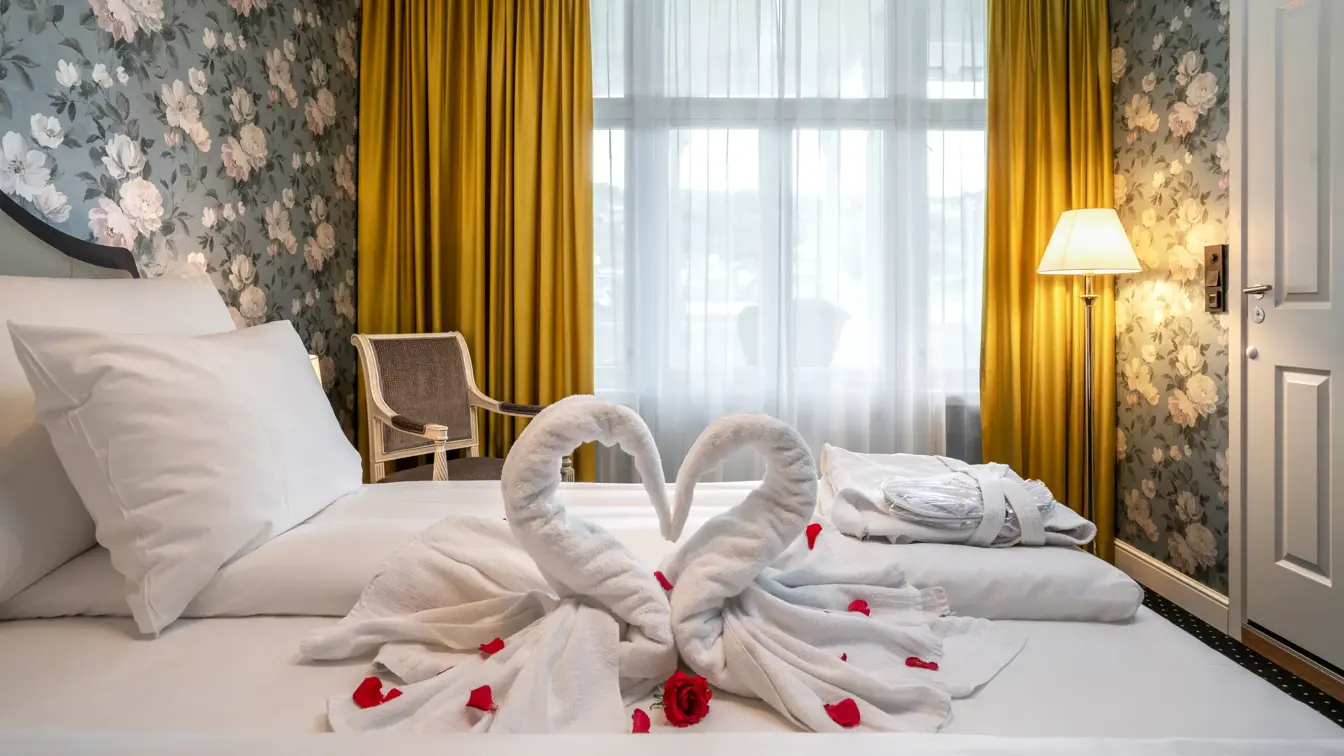 Dobbeltseng, svanehåndkle, rødes roseblader, morgenkåpevindu, gule gardiner, tapetvegg, stol, gulvlampe