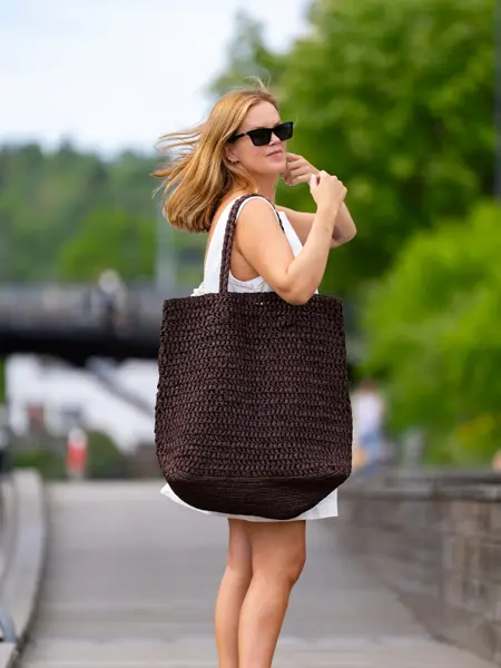 Dame i hvit kjole og sorte solbriller stå på en brygge med en stor brun flettet veske over skulderen.