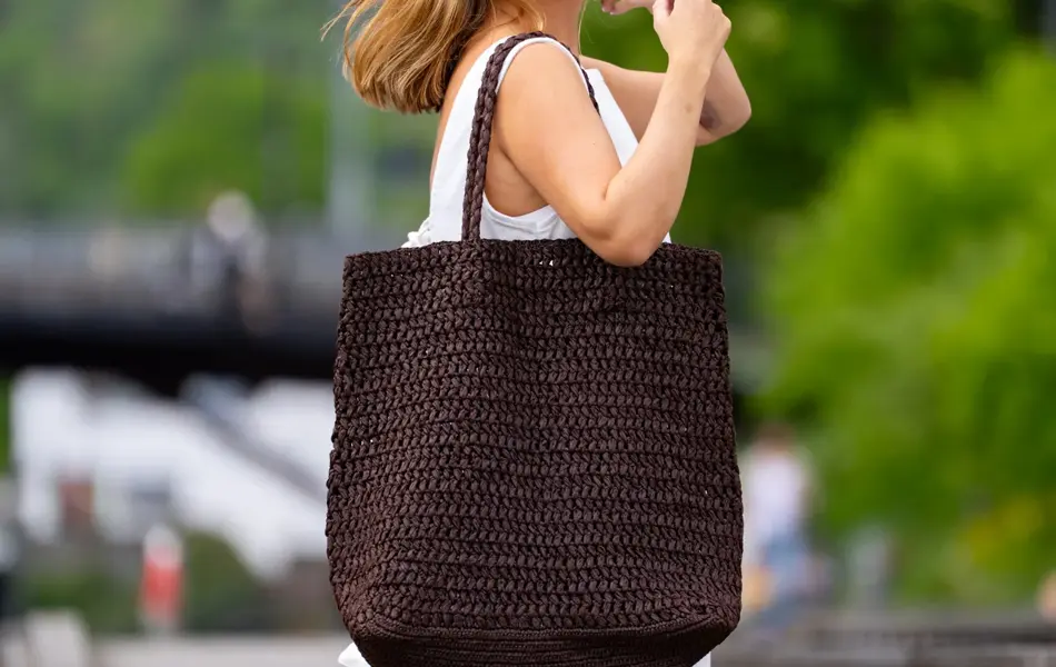 Dame i hvit kjole og sorte solbriller stå på en brygge med en stor brun flettet veske over skulderen.