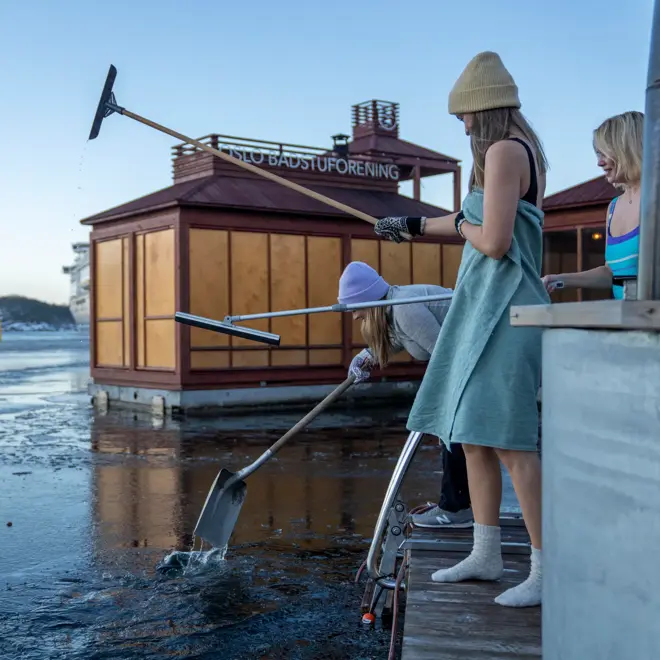 Badegjester i lue og håndkle hakker opp isen ved badetrappen foran en flytende badstue fra Oslo Badstuforening i Oslofjorden en vinterdag.