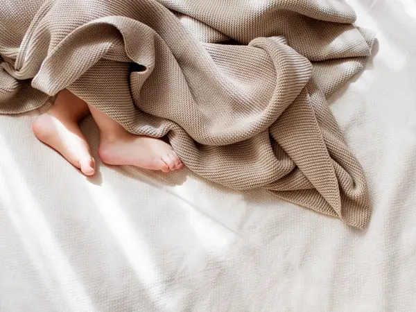 Baby som ligger i senga og sover meg brun strikket teppe over seg