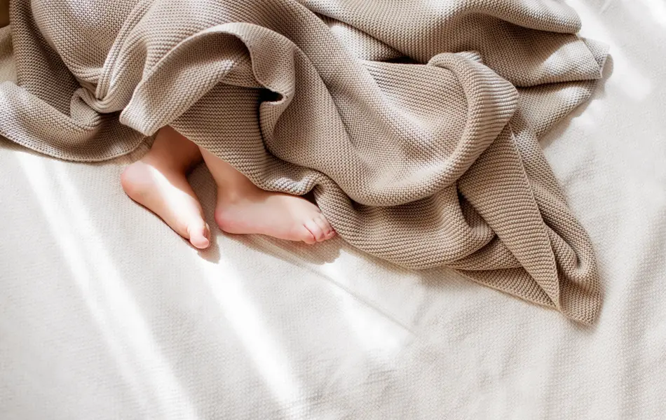 Baby som ligger i senga og sover meg brun strikket teppe over seg