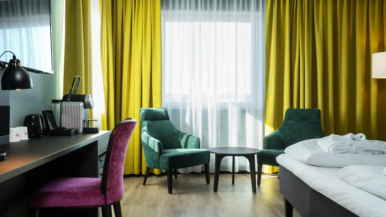 Salongbord, to grønne lenestoler, dobbeltseng, rosa lenestol, skrivebord, vindu, gule gardiner, TV 