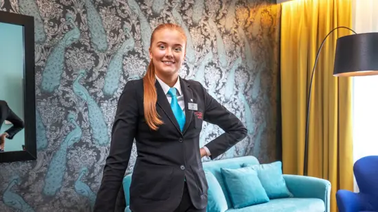 En kvinnelig ansatt i uniform, tapetvegg med mønster, gul gardin, turkis sofa, blå stol og et svart rundt bord. 