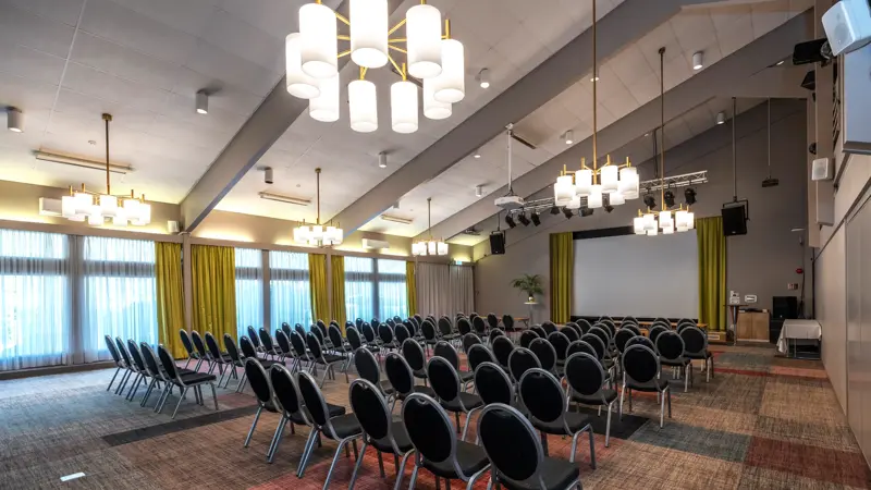 Stor sal med kino oppsett, lysekroner i taket, store vinduer med gule gardiner, stort lerret foran i lokalet