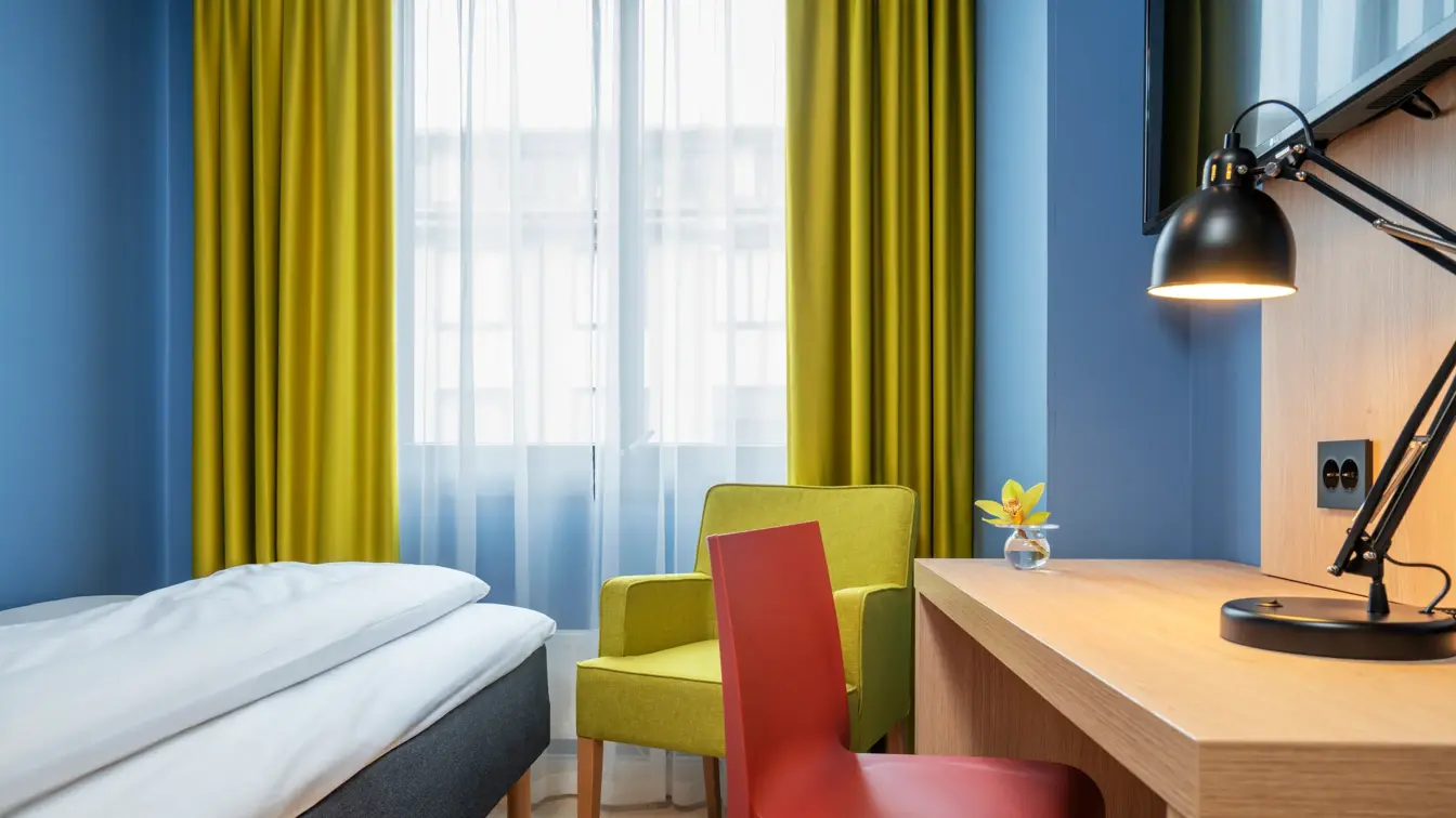 Enkelt seng, blå vegger, gul stol og gule gardiner, skrivebord og TV på enkeltrom på Thon Hotel Astoria