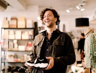 Mann på shopping med mørkt krøllete hår og brun jakke holder en sko i hendene inne i en klesbutikk