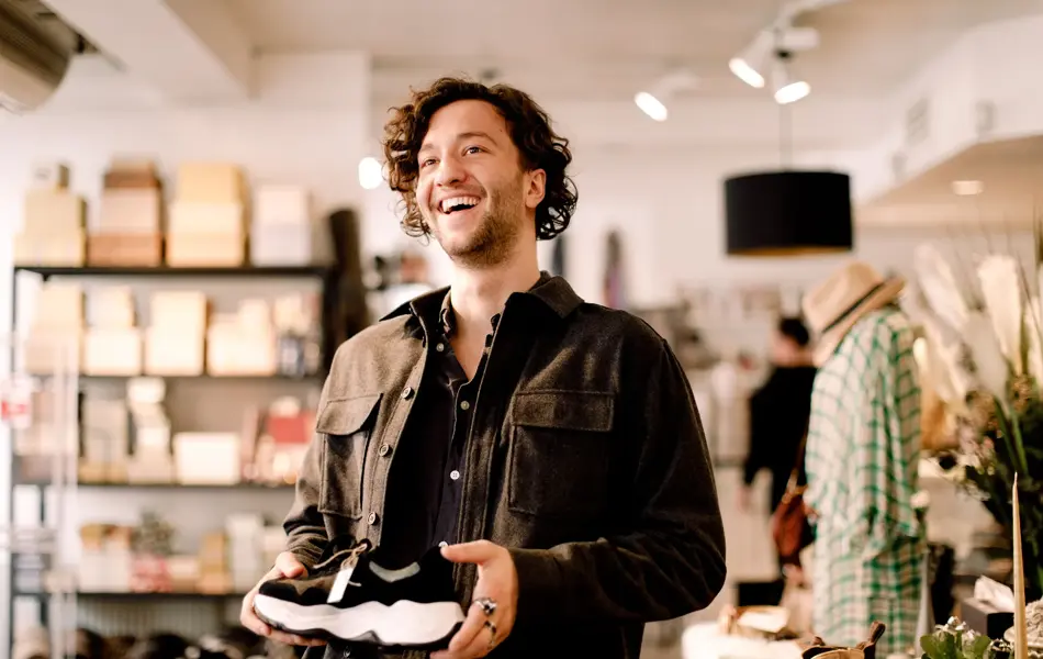 Mann på shopping med mørkt krøllete hår og brun jakke holder en sko i hendene inne i en klesbutikk
