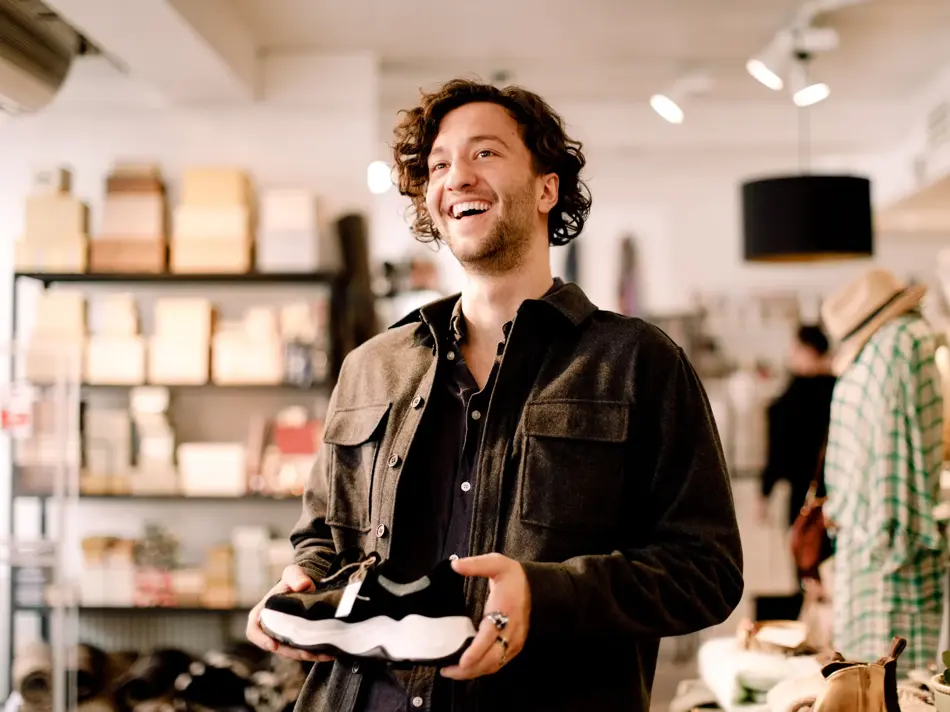 Mann på shopping med mørkt krøllete hår og brun jakke holder en sko i hendene inne i en klesbutikk