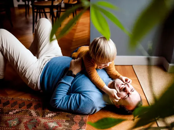 Mann ligger på gulvet og leker med et barn som er rundt to år, grønne toner i vegg og klær, gulvteppe i varme toner, en plante i uskarp forgrunn. Foto til artikkel om farsdagsgaver fra de minste barna.
