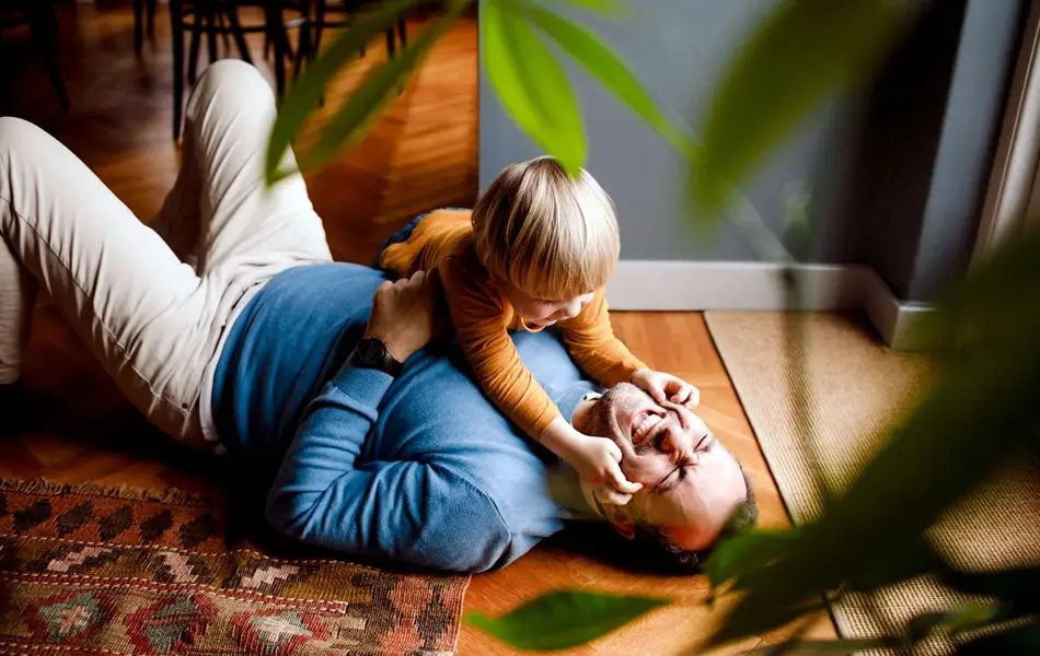 Mann ligger på gulvet og leker med et barn som er rundt to år, grønne toner i vegg og klær, gulvteppe i varme toner, en plante i uskarp forgrunn. Foto til artikkel om farsdagsgaver fra de minste barna.