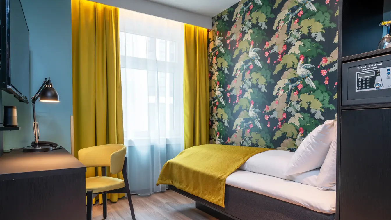 Skrivebord med leselampe og gul stol. Turkise vegger, vindu og gule gardiner. Fargerik tapet med fugler og blomster og enkeltseng i enkeltrom på Thon Hotel Bristol Bergen
