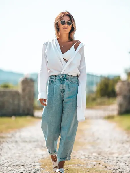 Dame går på grusvei iført hvit åpen bluse, hvit bh, sandaler, solbriller og løse baggy jeans.